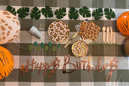 Birthday table setup with animal print plates, cutlery, and 'Happy Birthday' sign on a checkered tablecloth.