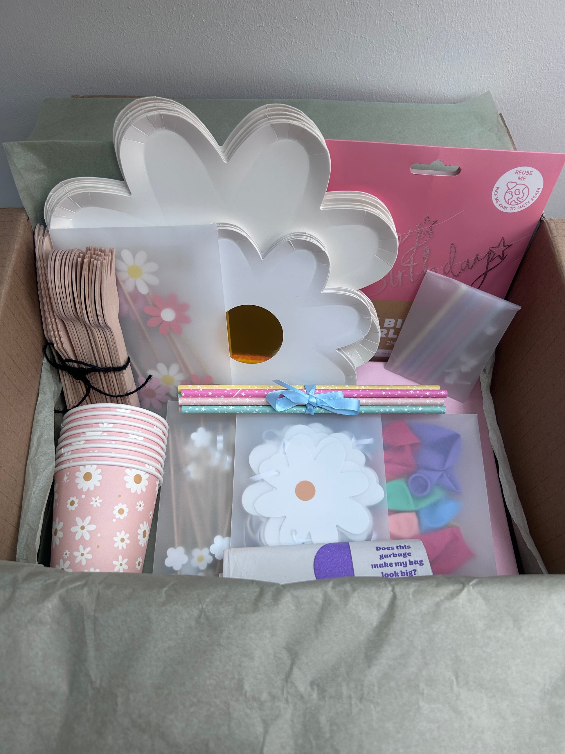 Assorted party supplies including a white flower-shaped cake stand, pink cups with floral designs, and other decorative items on a gray surface.