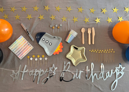 Birthday party supplies including balloons, plates, cutlery, and 'Happy Birthday' banner on a gray surface.