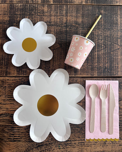 White daisy-shaped plates with gold centers on a wooden surface, accompanied by a pink paper cup and utensils.
